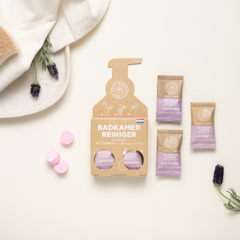 Eco-friendly lavender bathroom cleaner refill set by The Green Lab Co., featuring a cardboard package shaped like a spray bottle with three pink tablets inside. Three individually wrapped lavender-scented tablets are shown next to the box. Surrounding items include a white towel, lavender sprigs, and a natural sponge on a cream-colored background.